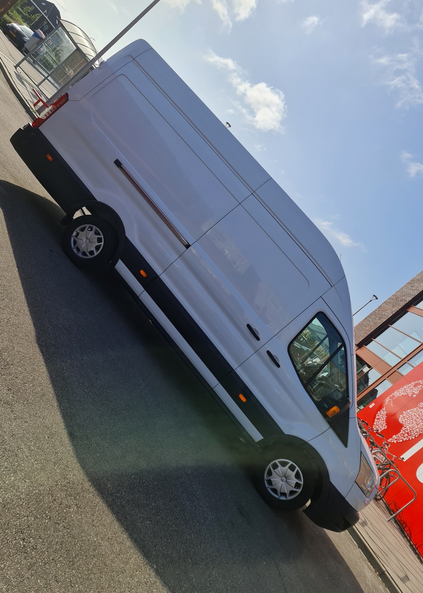 Biały van dostawczy zaparkowany na parkingu, widoczne odbicie w szybie, budynek z czerwonym elementem dekoracyjnym w tle, jasne niebo.