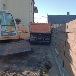 PIOTRO-TRANS Piotr Ostasiewicz - Pomarańczowa koparka i wywrotka Volvo na placu budowy między budynkami. Widoczny fragment drewnianego płotu i ślady opon na ziemi. Prace ziemne.