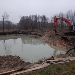 PIOTRO-TRANS Piotr Ostasiewicz - Pomarańczowa koparka Hitachi przy wykopanym stawie, w tle drzewa bez liści i pochmurne niebo. Widoczna pompa do wody i węże. Prace ziemne w toku.