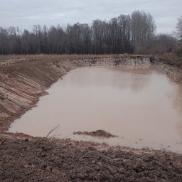 PIOTRO-TRANS Piotr Ostasiewicz - Wykonany wykop pod zbiornik wodny wypełniony wodą, z widocznymi śladami maszyn na brzegach. W tle drzewa bez liści i pochmurne niebo.