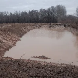 Wykonany wykop pod zbiornik wodny wypełniony wodą, z widocznymi śladami maszyn na brzegach. W tle drzewa bez liści i pochmurne niebo.