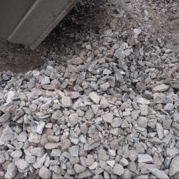 PIOTRO-TRANS Piotr Ostasiewicz - Kruszywo skalne w odcieniach szarości i beżu, częściowo przesypane przez łyżkę koparki. Widoczna frakcja kamieni o różnej wielkości, tło z ciemniejszej ziemi.