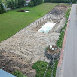 PIOTRO-TRANS Piotr Ostasiewicz - Widok z góry na wykop pod zbiornik wodny, zabezpieczony palikami i taśmą. Obok droga, pole i gniazdo bocianów na słupie. Ślady opon na ziemi wskazują na prace ziemne.