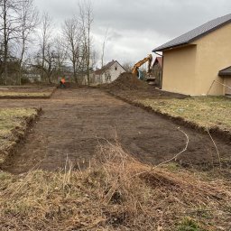 PIOTRO-TRANS Piotr Ostasiewicz - Przygotowany teren pod budowę z wykopanymi fundamentami, koparka w tle, pracownik w pomarańczowej kamizelce. Widoczne budynki w otoczeniu.