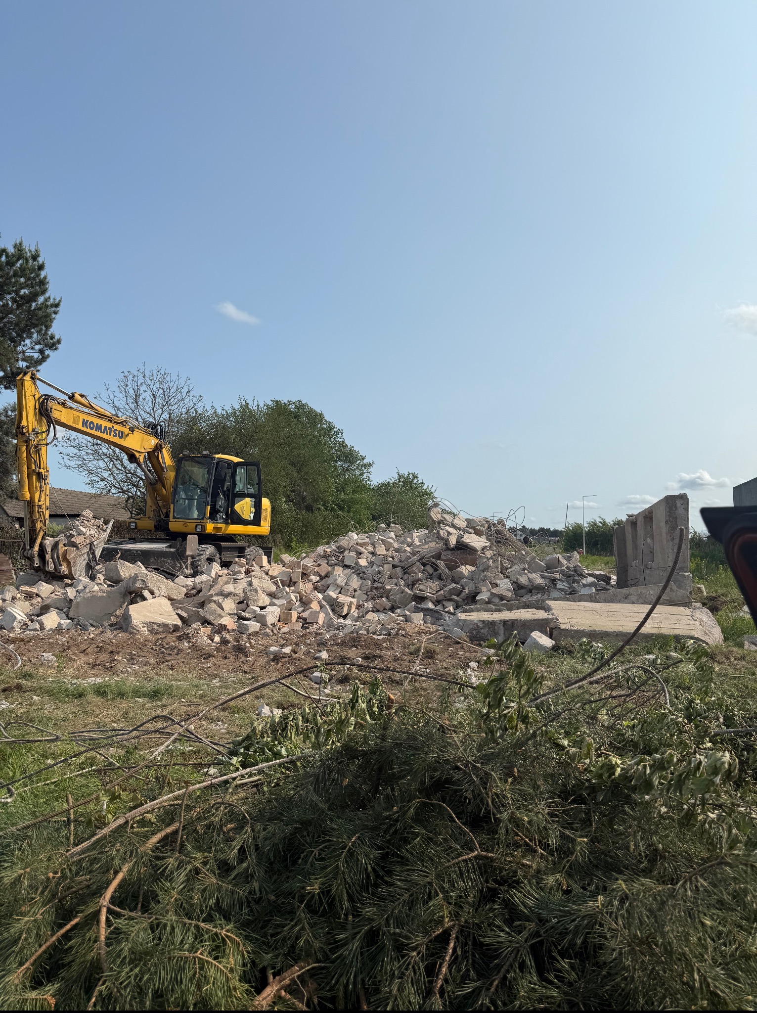Żółta koparka Komatsu na placu budowy, usuwająca pozostałości po rozbiórce budynku. Na pierwszym planie gałęzie drzew iglastych. Błękitne niebo w tle.