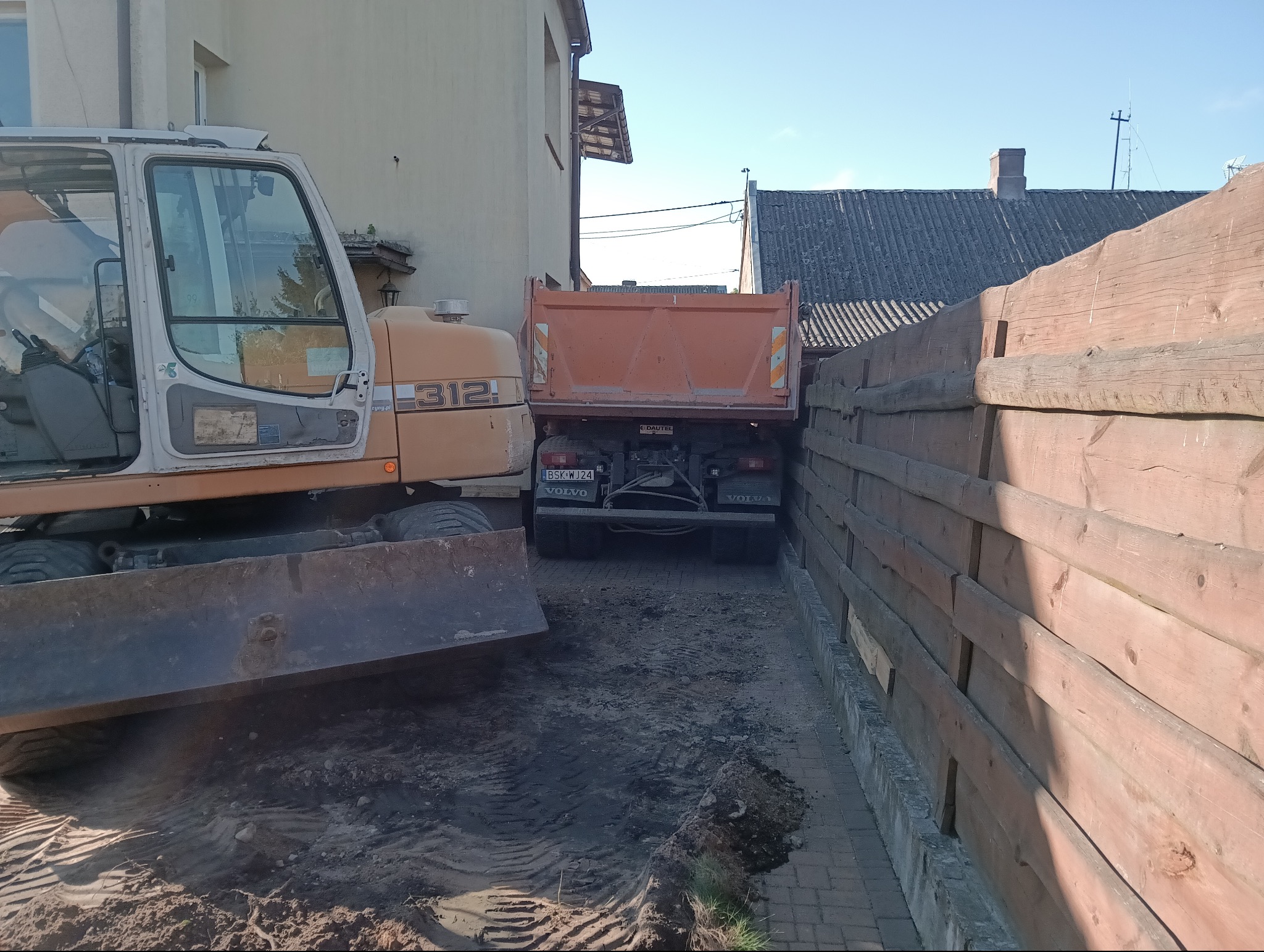 Pomarańczowa koparka i wywrotka Volvo na placu budowy między budynkami. Widoczny fragment drewnianego płotu i ślady opon na ziemi. Prace ziemne.