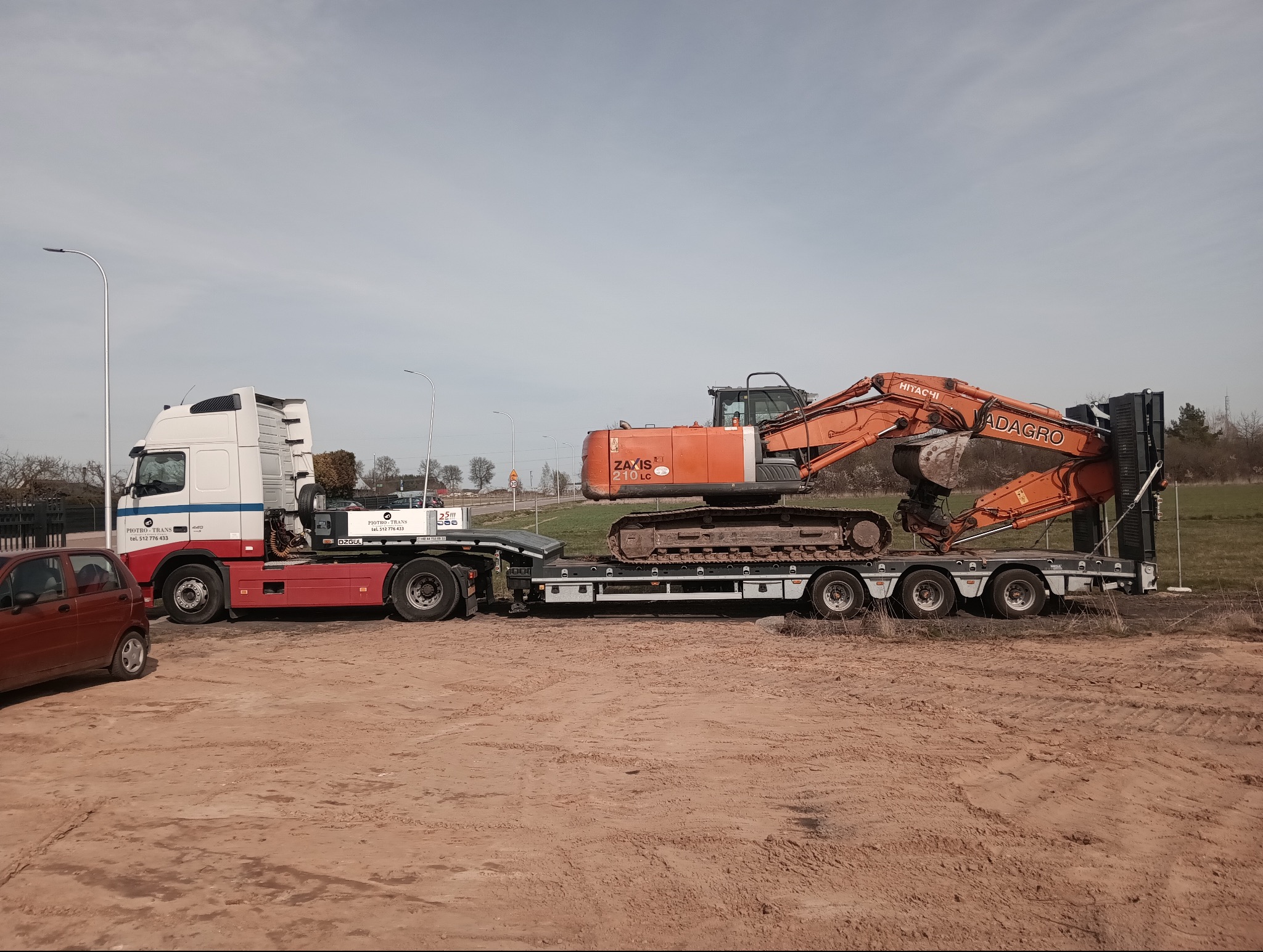 Pomarańczowa koparka Hitachi ZAXY 210 LC na niskopodwoziowej naczepie ciągniętej przez biało-czerwony ciągnik siodłowy firmy Piotro-Trans, transport sprzętu budowlanego.
