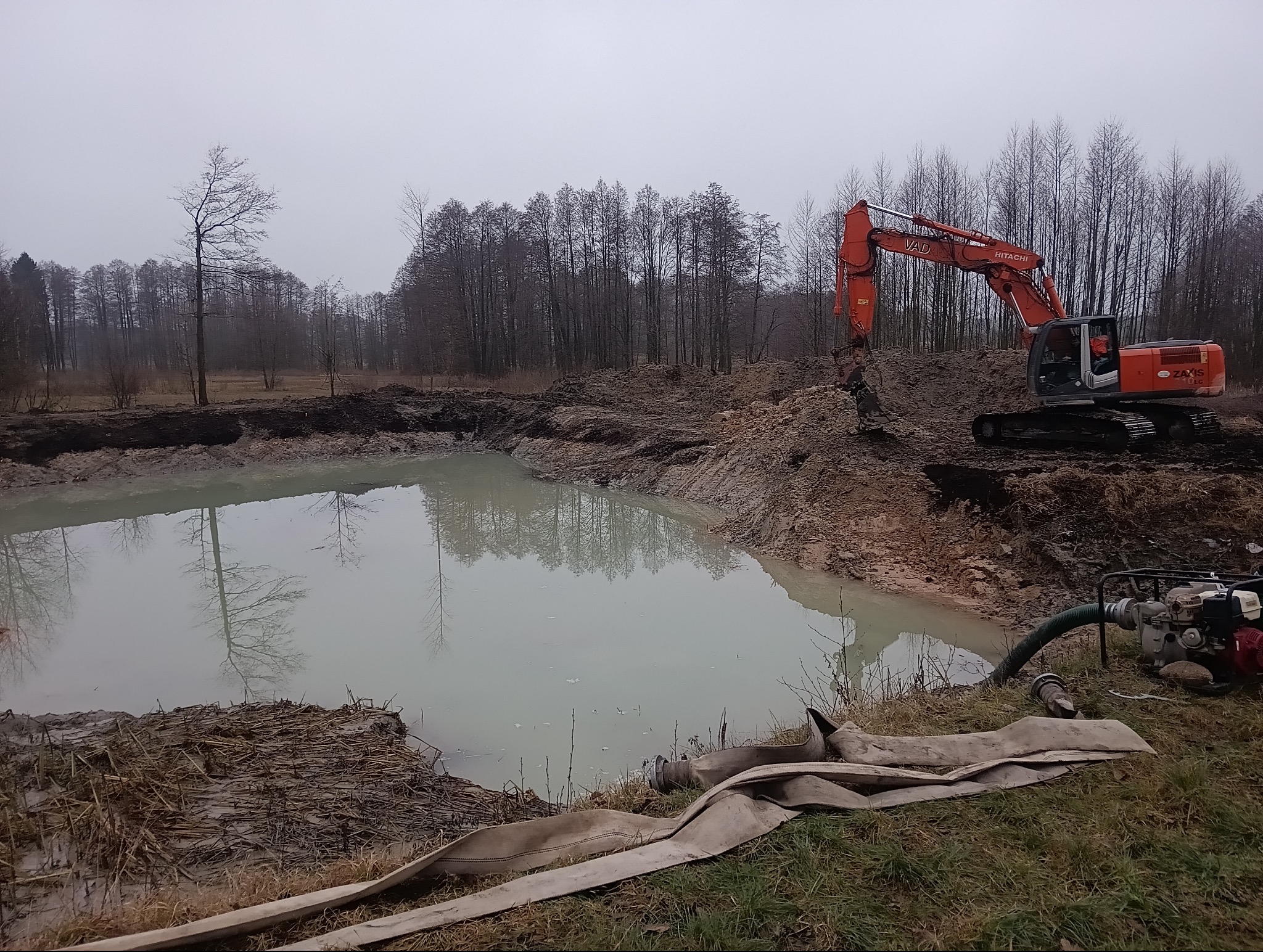 Pomarańczowa koparka Hitachi przy wykopanym stawie, w tle drzewa bez liści i pochmurne niebo. Widoczna pompa do wody i węże. Prace ziemne w toku.