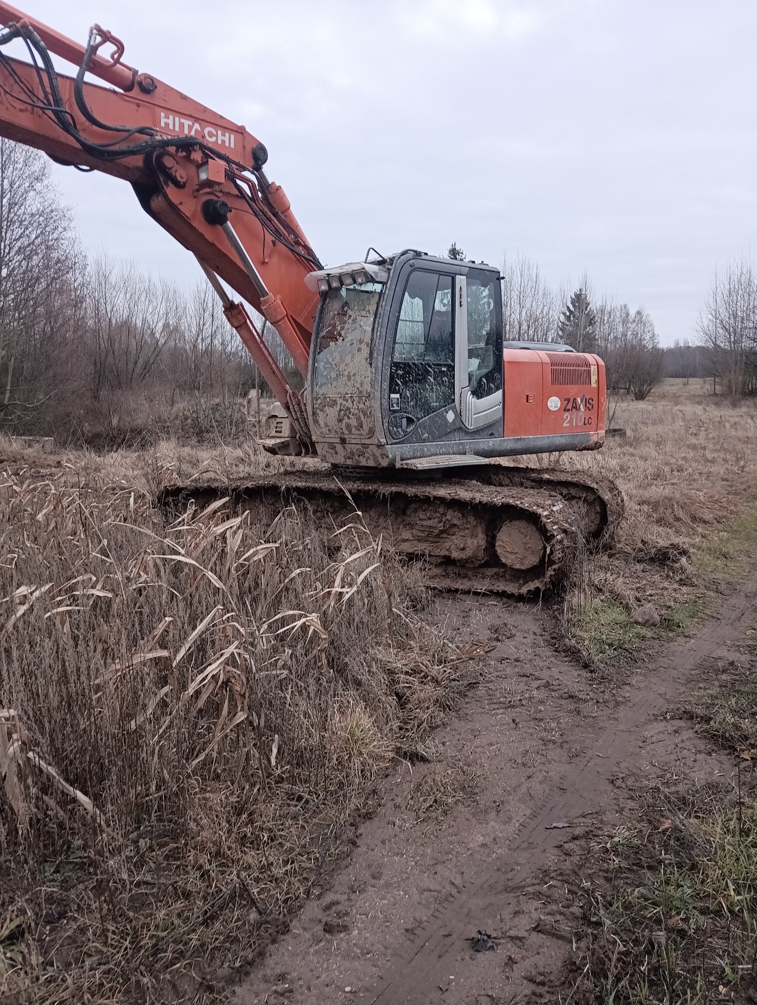 Pomarańczowa koparka Hitachi Zaxis 210LC w błotnistym terenie z suchą trawą, po wykonanych pracach. Kabina ubrudzona błotem, pochmurne niebo w tle.