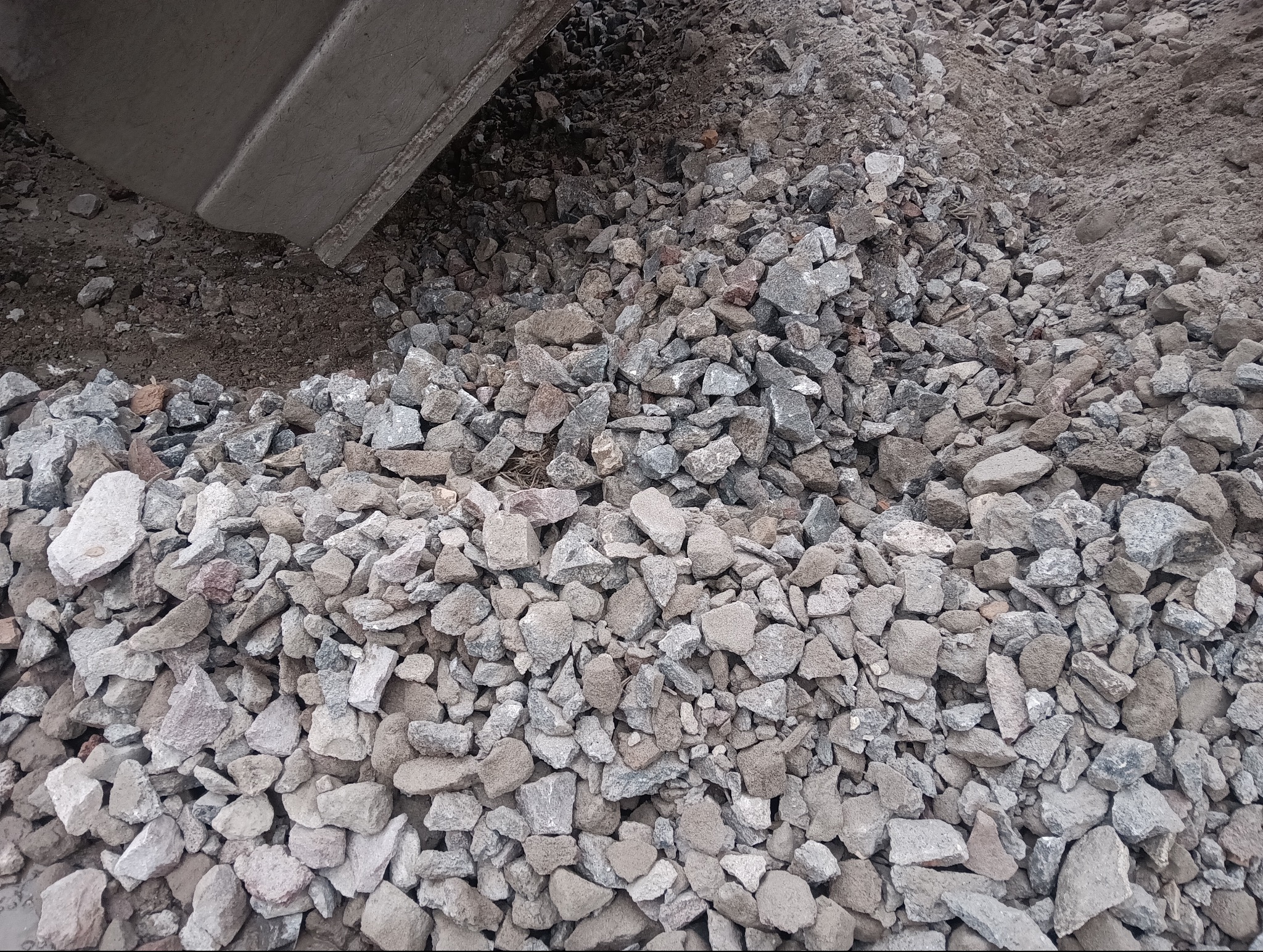 Kruszywo skalne w odcieniach szarości i beżu, częściowo przesypane przez łyżkę koparki. Widoczna frakcja kamieni o różnej wielkości, tło z ciemniejszej ziemi.