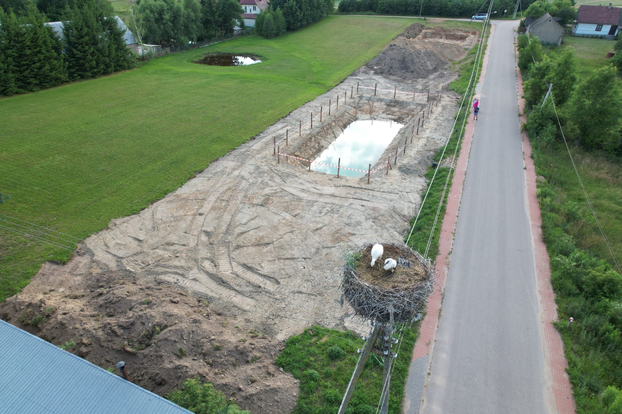 Widok z góry na wykop pod zbiornik wodny, zabezpieczony palikami i taśmą. Obok droga, pole i gniazdo bocianów na słupie. Ślady opon na ziemi wskazują na prace ziemne.