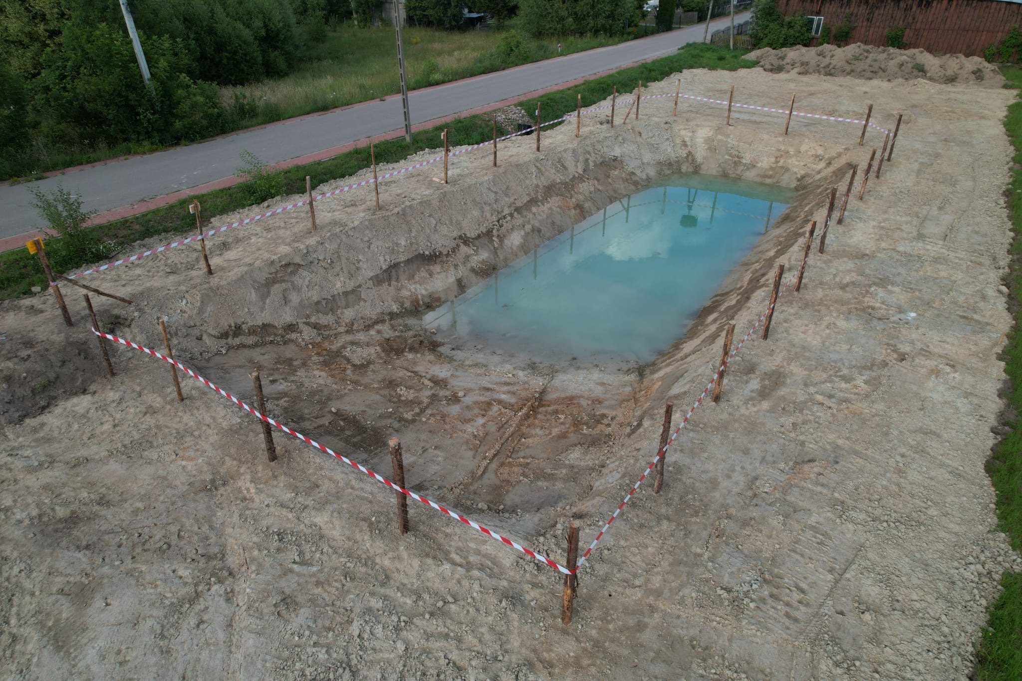 Wykopany dół wypełniony wodą, zabezpieczony palikami i taśmą ostrzegawczą. Widok z góry na wykop w ziemi, w tle droga i zieleń.