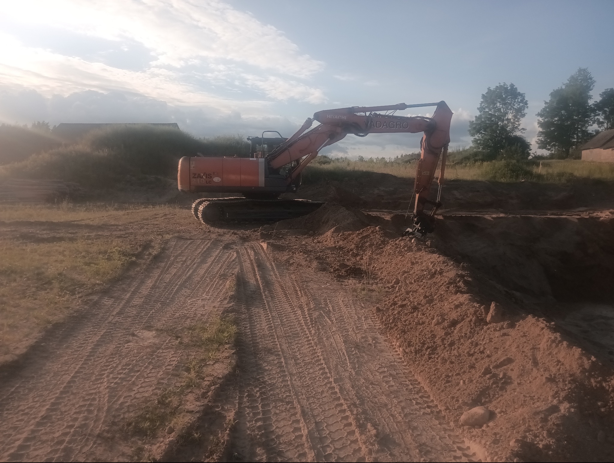 Pomarańczowa koparka Hitachi Vadagro w trakcie prac ziemnych, widoczne ślady opon na gruncie i wykop. W tle zabudowania i błękitne niebo.