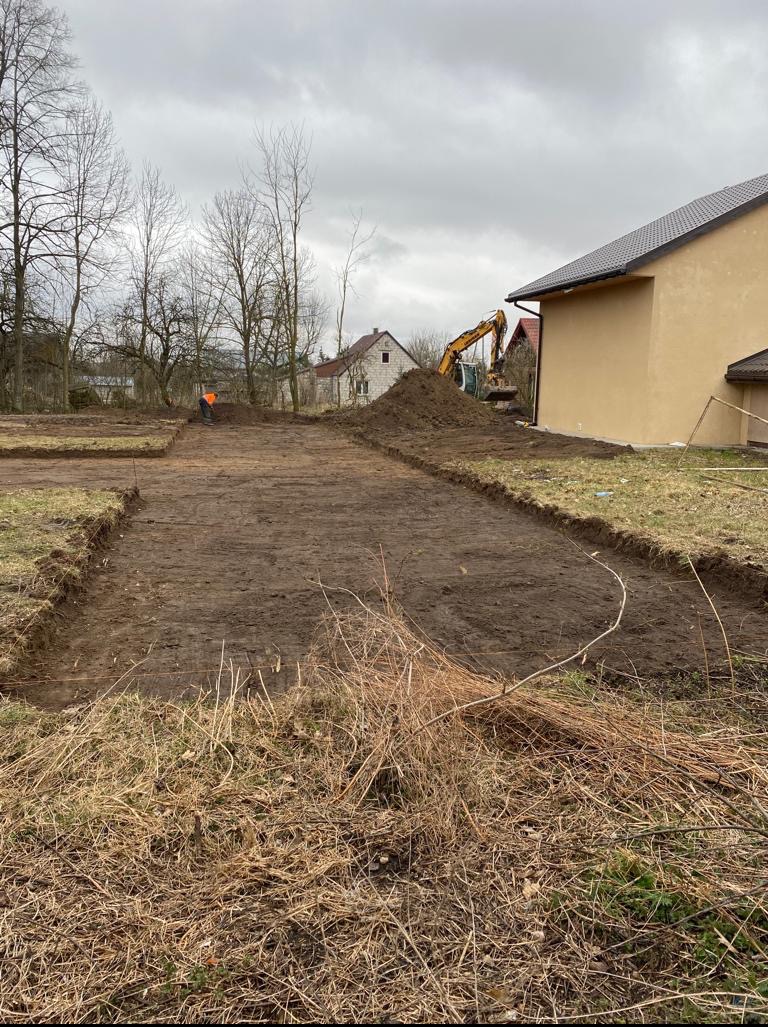 Przygotowany teren pod budowę z wykopanymi fundamentami, koparka w tle, pracownik w pomarańczowej kamizelce. Widoczne budynki w otoczeniu.