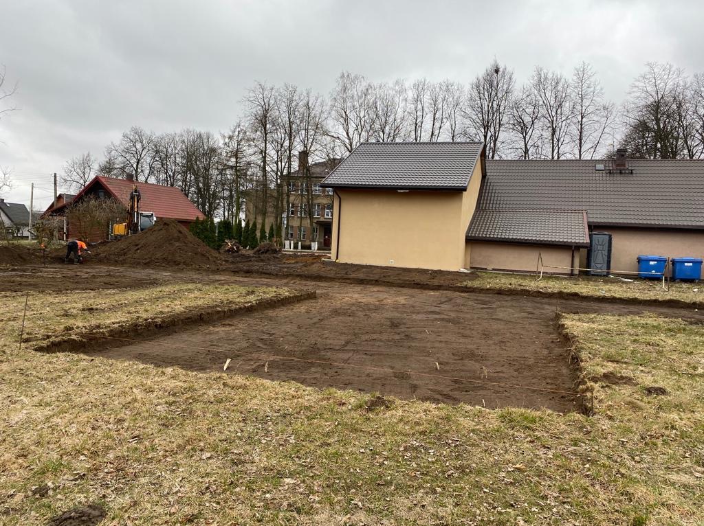 Wykopane fundamenty pod budynek, widoczny wykop, pracownik i koparka w tle, pochmurne niebo.