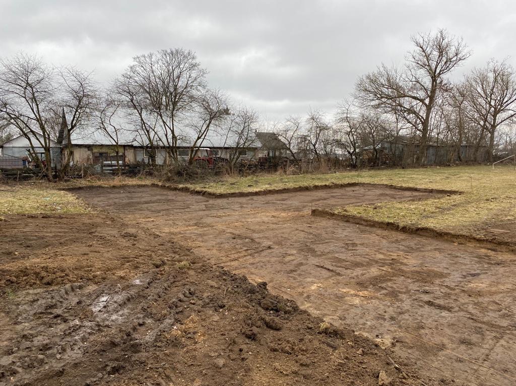 Przygotowany, wilgotny teren pod budowę z widocznymi wykopami pod fundamenty. W tle budynki gospodarcze i drzewa bez liści pod pochmurnym niebem.