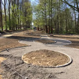 Ścieżka parkowa w trakcie budowy z wyprofilowanymi rabatami, widoczna ubijarka wibracyjna i narzędzia.