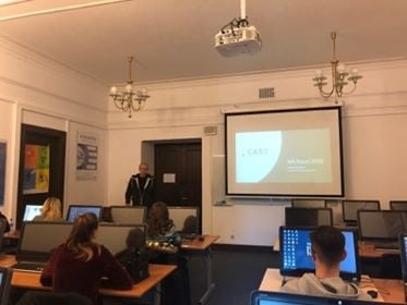 Sala komputerowa z rzędami stanowisk, studenci siedzą przed monitorami, prowadzący stoi przy ekranie projekcyjnym z prezentacją, na ścianach widać zabytkowe lampy i elementy dekoracyjne.