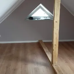 Poddasze z drewnianą konstrukcją wspierającą, triangularne okno dachowe i panele podłogowe imitujące drewno.