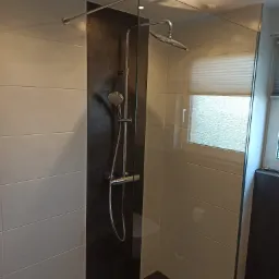 Nowoczesna kabina prysznicowa typu walk-in z szklaną ścianką, chromowaną armaturą i płytkami w odcieniach bieli i grafitu, widok na okno z żaluzjami.