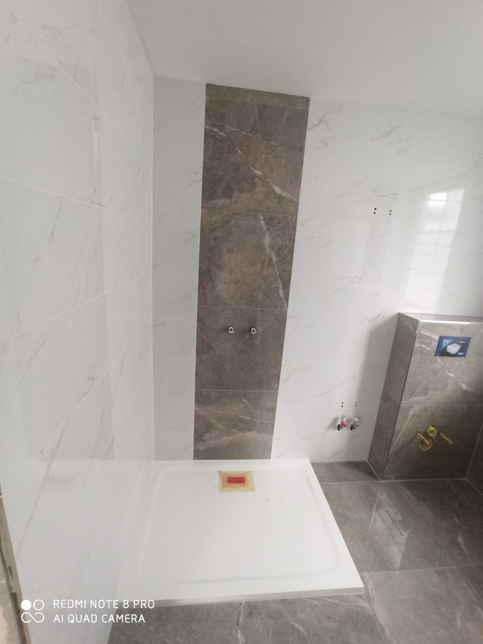 Wnętrze łazienki w trakcie wykańczania, widoczny brodzik, płytki imitujące marmur na ścianach i podłodze, oraz elementy instalacji wodnej.