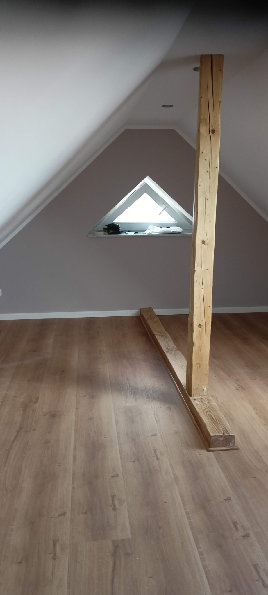 Poddasze z drewnianą konstrukcją wspierającą, triangularne okno dachowe i panele podłogowe imitujące drewno.