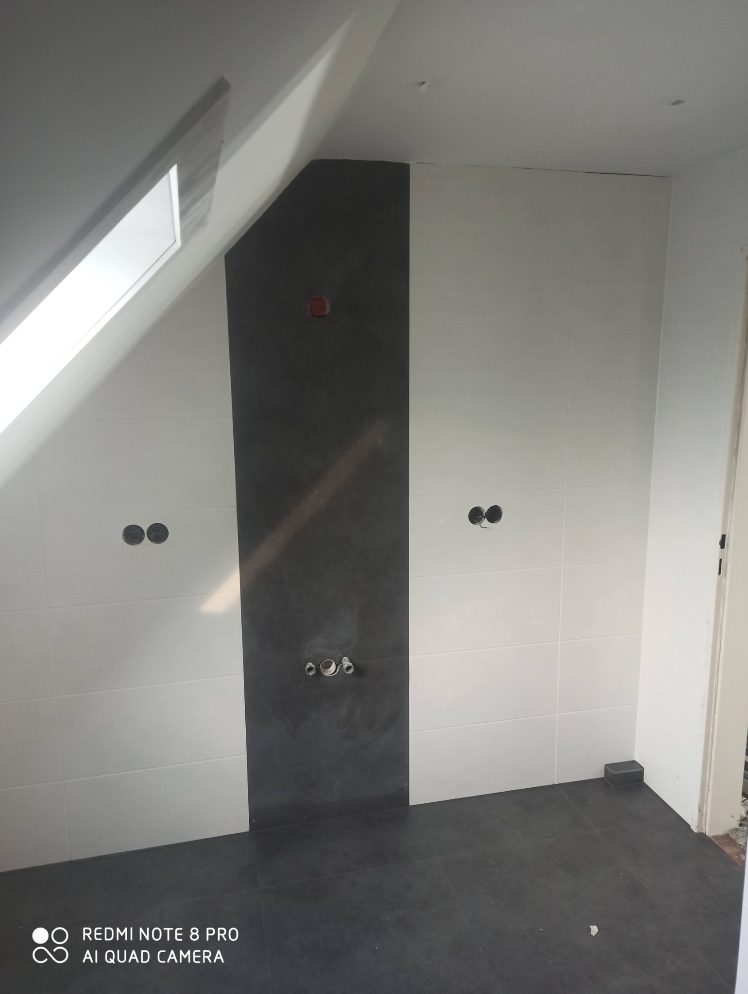 Ukończona łazienka na poddaszu ze skośnym oknem dachowym, ściany wyłożone białymi kafelkami z ciemnoszarym pasem na środku, widoczne otwory na baterie i rury, ciemna podłoga z płytek.