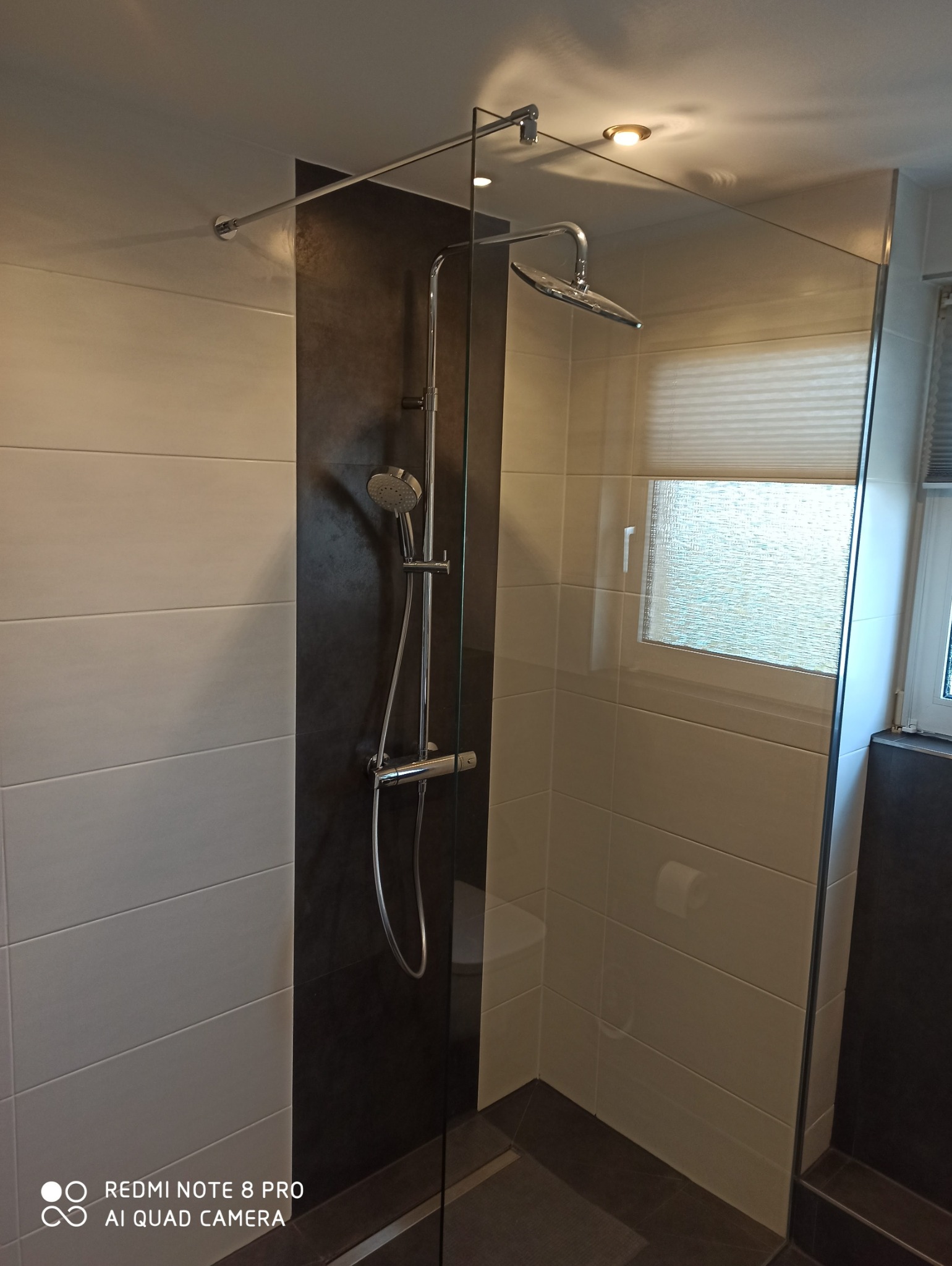 Nowoczesna kabina prysznicowa typu walk-in z szklaną ścianką, chromowaną armaturą i płytkami w odcieniach bieli i grafitu, widok na okno z żaluzjami.