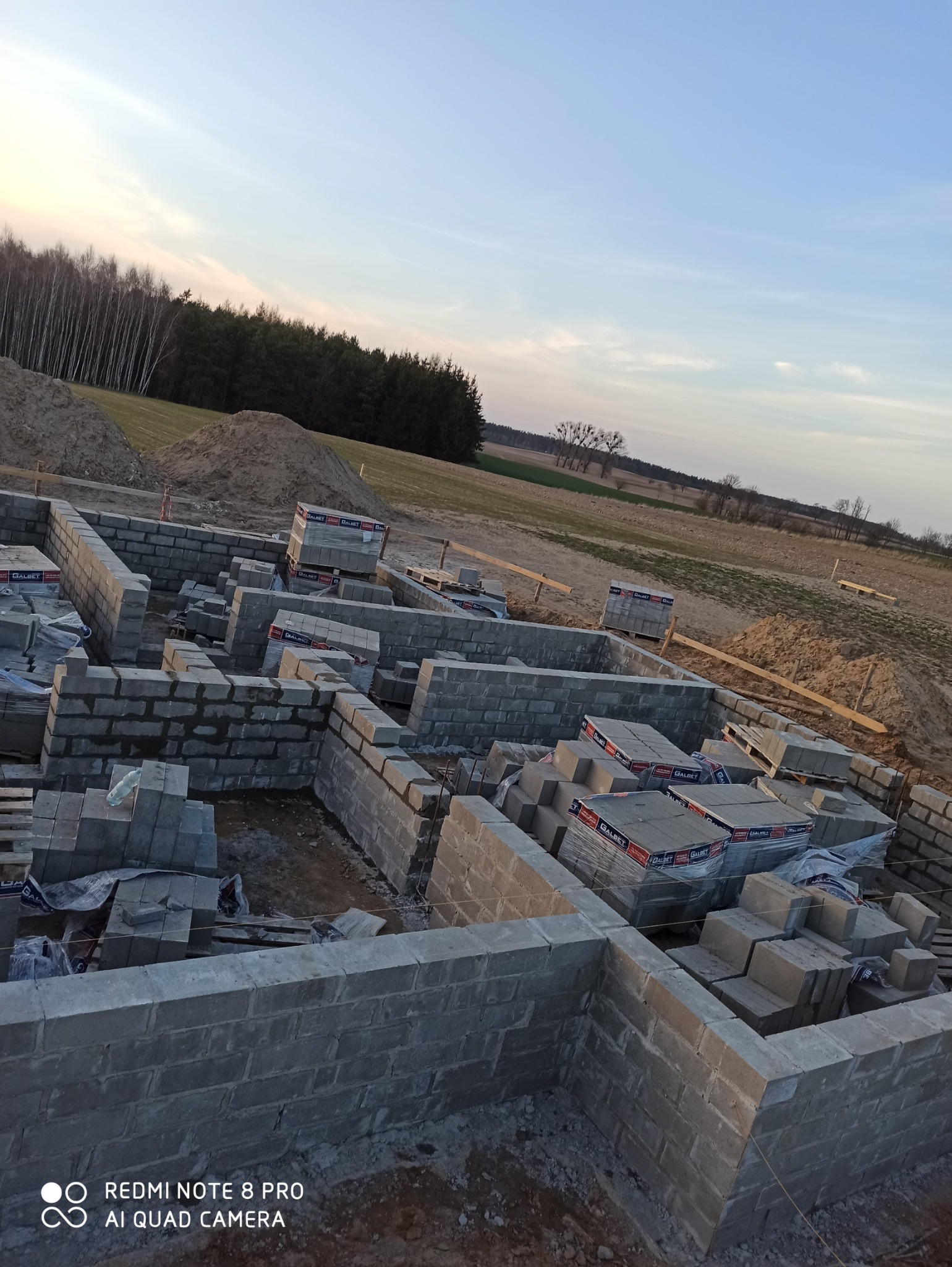 Fundamenty domu w trakcie budowy, w tle widoczne pole i las, palety z bloczkami czekają na użycie.