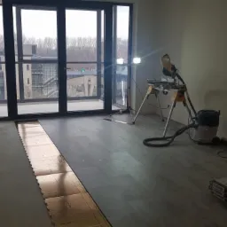 Układanie paneli podłogowych w jasnym pokoju z dużym oknem balkonowym, widoczna piła elektryczna, odkurzacz przemysłowy i paczki paneli.