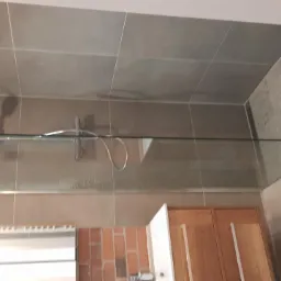 Szklana kabina prysznicowa typu walk-in z deszczownicą zamontowaną na ścianie wyłożonej szarymi, prostokątnymi płytkami; widoczna część szafki pod umywalkę z drewnianymi frontami oraz fragment...