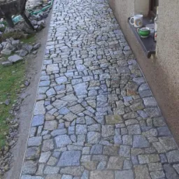 Ułożona ścieżka z nieregularnych kamieni brukowych w odcieniach szarości i beżu, prowadząca wzdłuż ściany budynku z widocznym oknem i zielonym wężem ogrodowym, z taczką i stertą kamieni w tle.