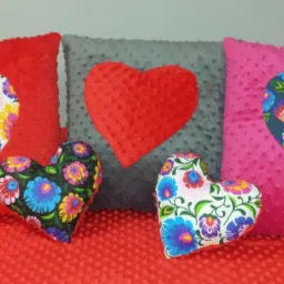 Dekoracyjne poduszki w kształcie serc i kwadratowe, w kolorach czerwonym, szarym i różowym, z aplikacjami w kwiatowe wzory, ułożone na czerwonej, teksturowanej powierzchni.