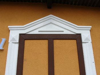 Detal elewacji budynku z dekoracyjną sztukaterią: biały naczółek trójkątny nad brązową ramą okienną, zakończoną elementami w kształcie ostrosłupów. Tło stanowi pomarańczowy tynk.