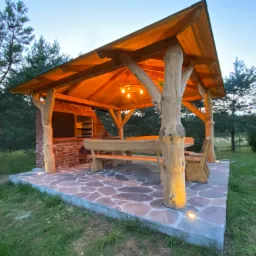 Drewniana pergola z oświetleniem, stołem i ławkami na kamiennej posadzce, obok murowany grill, oświetlona wieczorem.