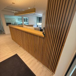 Rychert Holzdesign - Nowoczesna drewniana zabudowa lady recepcyjnej z pionowych listew w biurze. Widoczne oświetlenie sufitowe i fragment podłogi z ciemnym dywanem. Jasna kolorystyka wnętrza.