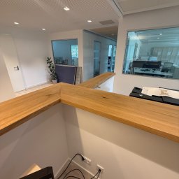 Rychert Holzdesign - Nowoczesne biuro z drewnianym blatem recepcyjnym, widok z góry na kable i gniazdka. Minimalistyczny design, jasne wnętrze z rośliną w tle i przeszklonymi ścianami.