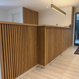 Rychert Holzdesign - Drewniana lamelowa zabudowa ściany i lady recepcyjnej z oświetleniem, widok z bliska, jasna podłoga, w tle drzwi i fragment wnętrza. Nowoczesny design.
