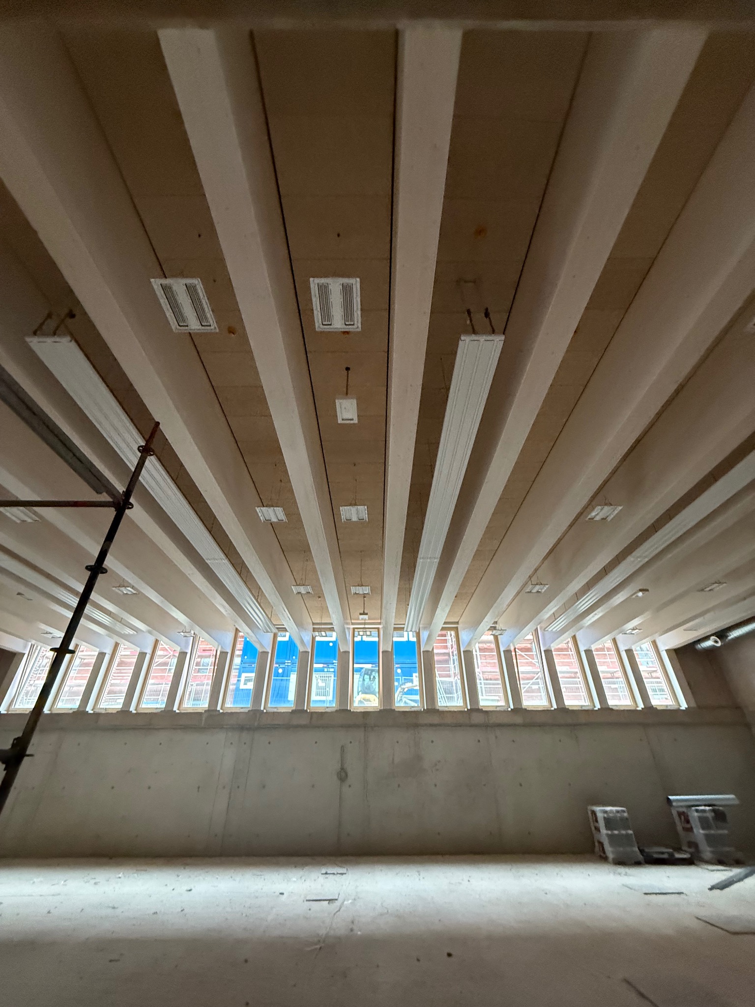 Wnętrze w trakcie budowy z drewnianym stropem belkowym i rzędem okien pod sufitem. Widoczne rusztowanie i materiały budowlane. Surowy stan wykończenia.