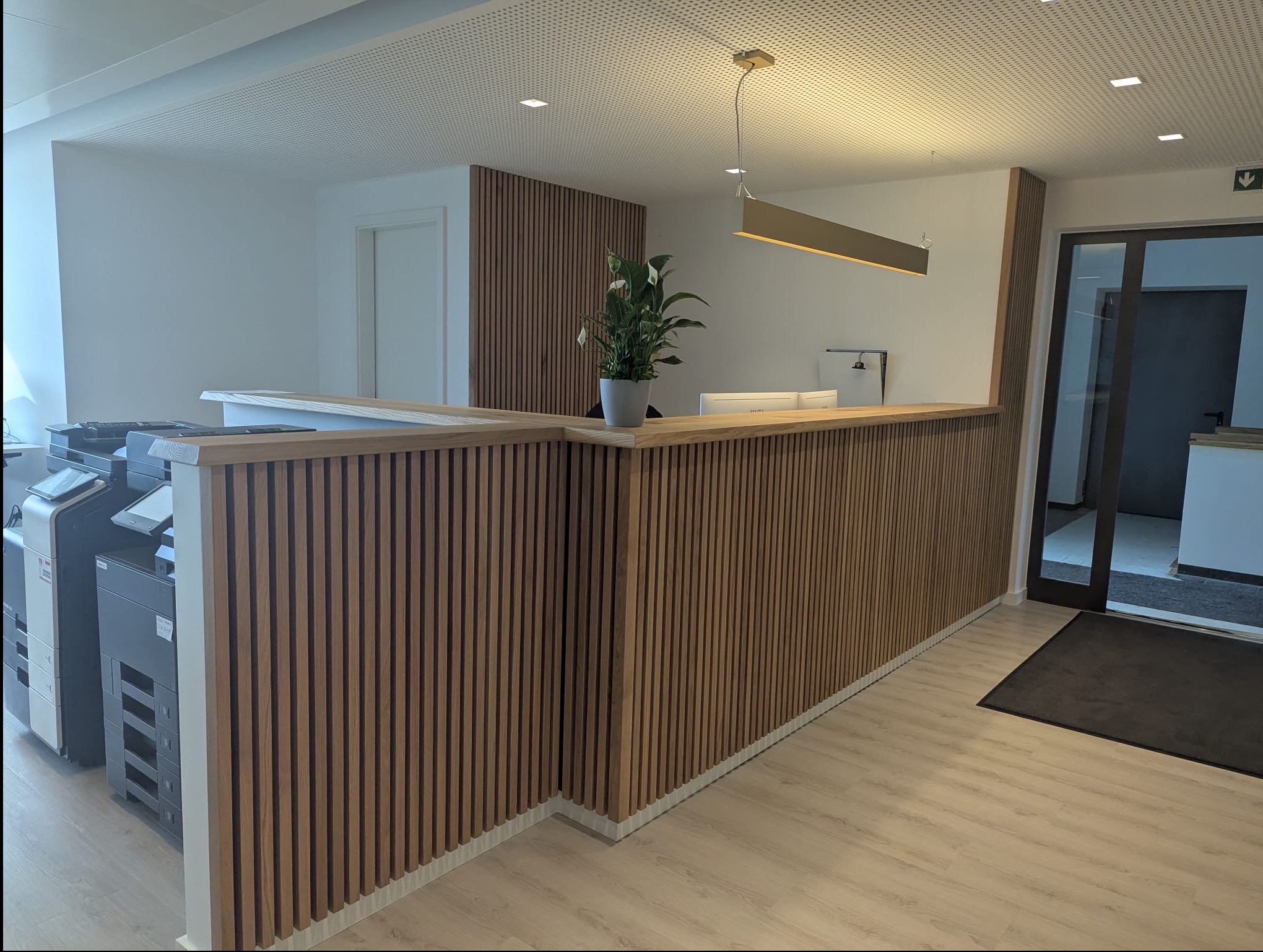 Nowoczesna lamelowa zabudowa recepcji z drewnianym blatem i rośliną w doniczce. W tle widoczne drzwi i elementy biurowe, oświetlenie sufitowe.