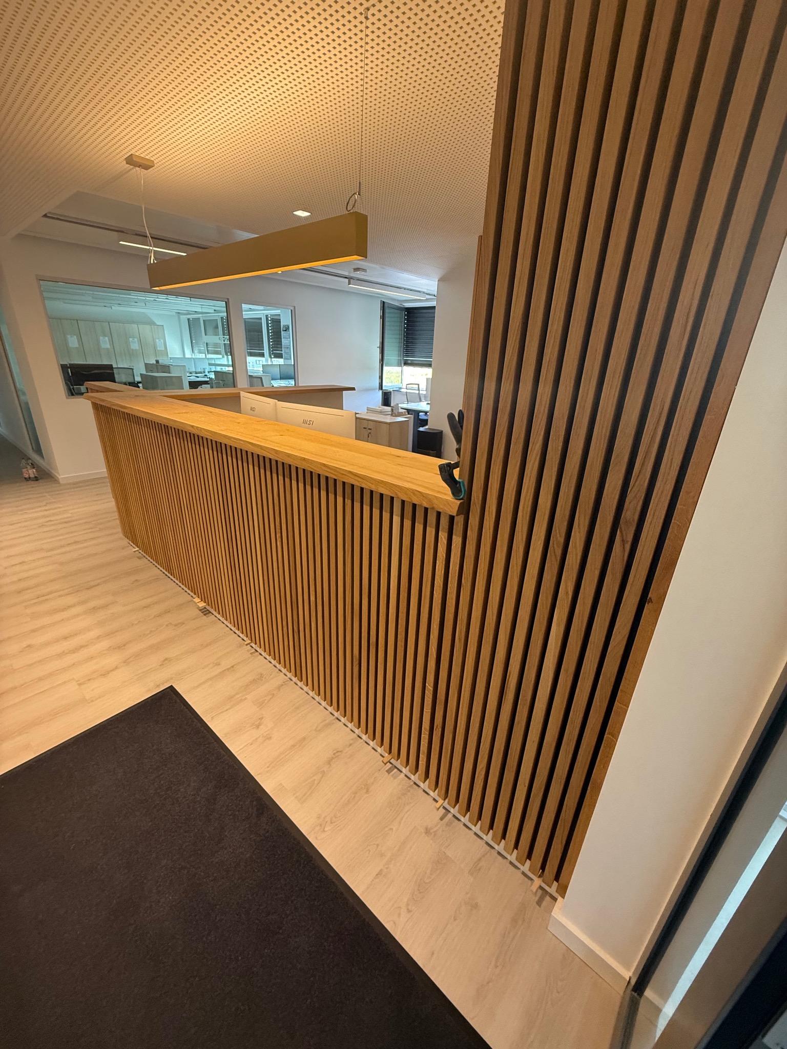 Nowoczesna drewniana zabudowa lady recepcyjnej z pionowych listew w biurze. Widoczne oświetlenie sufitowe i fragment podłogi z ciemnym dywanem. Jasna kolorystyka wnętrza.