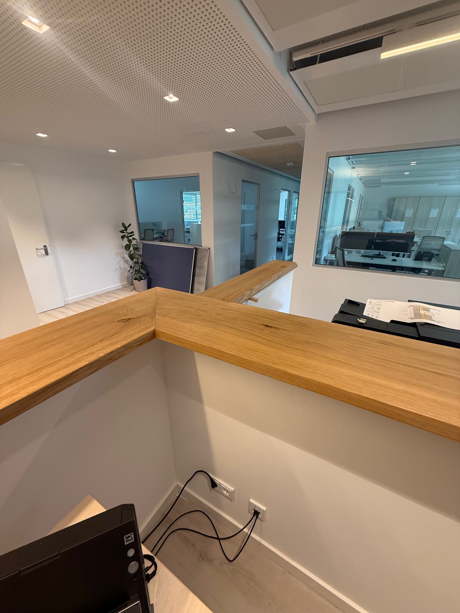 Nowoczesne biuro z drewnianym blatem recepcyjnym, widok z góry na kable i gniazdka. Minimalistyczny design, jasne wnętrze z rośliną w tle i przeszklonymi ścianami.