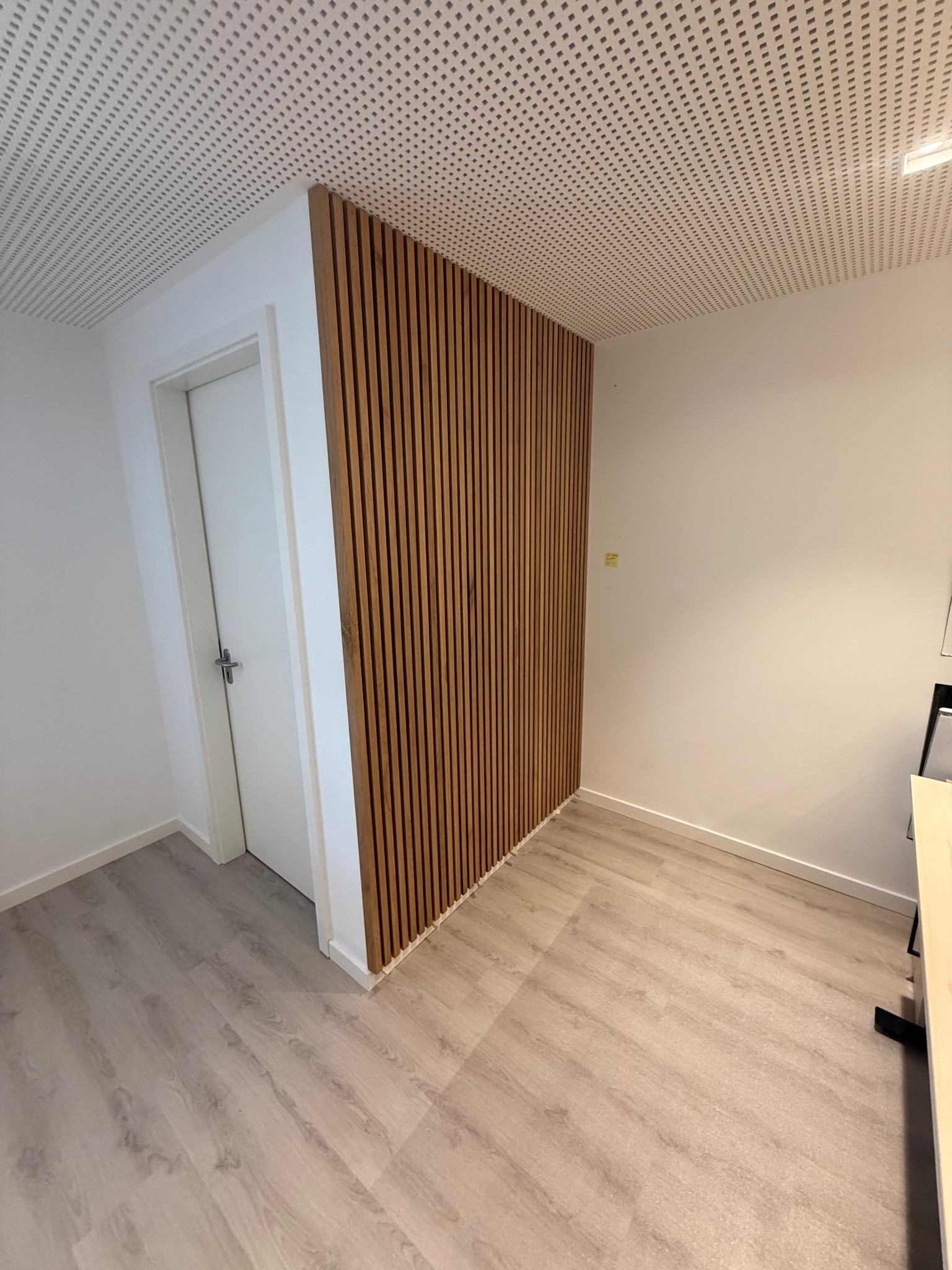 Drewniana ściana z paneli w wąskim korytarzu, z drzwiami po lewej i perforowanym sufitem. Jasne wnętrze z podłogą imitującą drewno.