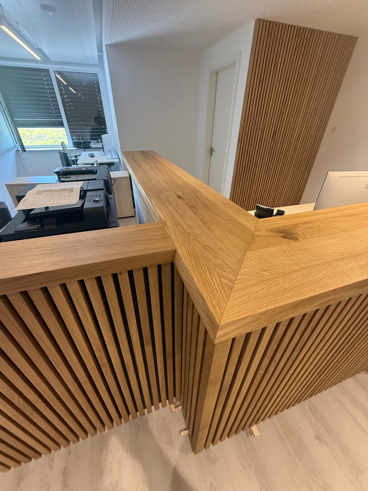 Drewniana lamelowa zabudowa biurowa z blatem w jasnym odcieniu drewna, widok z bliska na detal wykończenia. W tle biuro z oknem i żaluzjami.