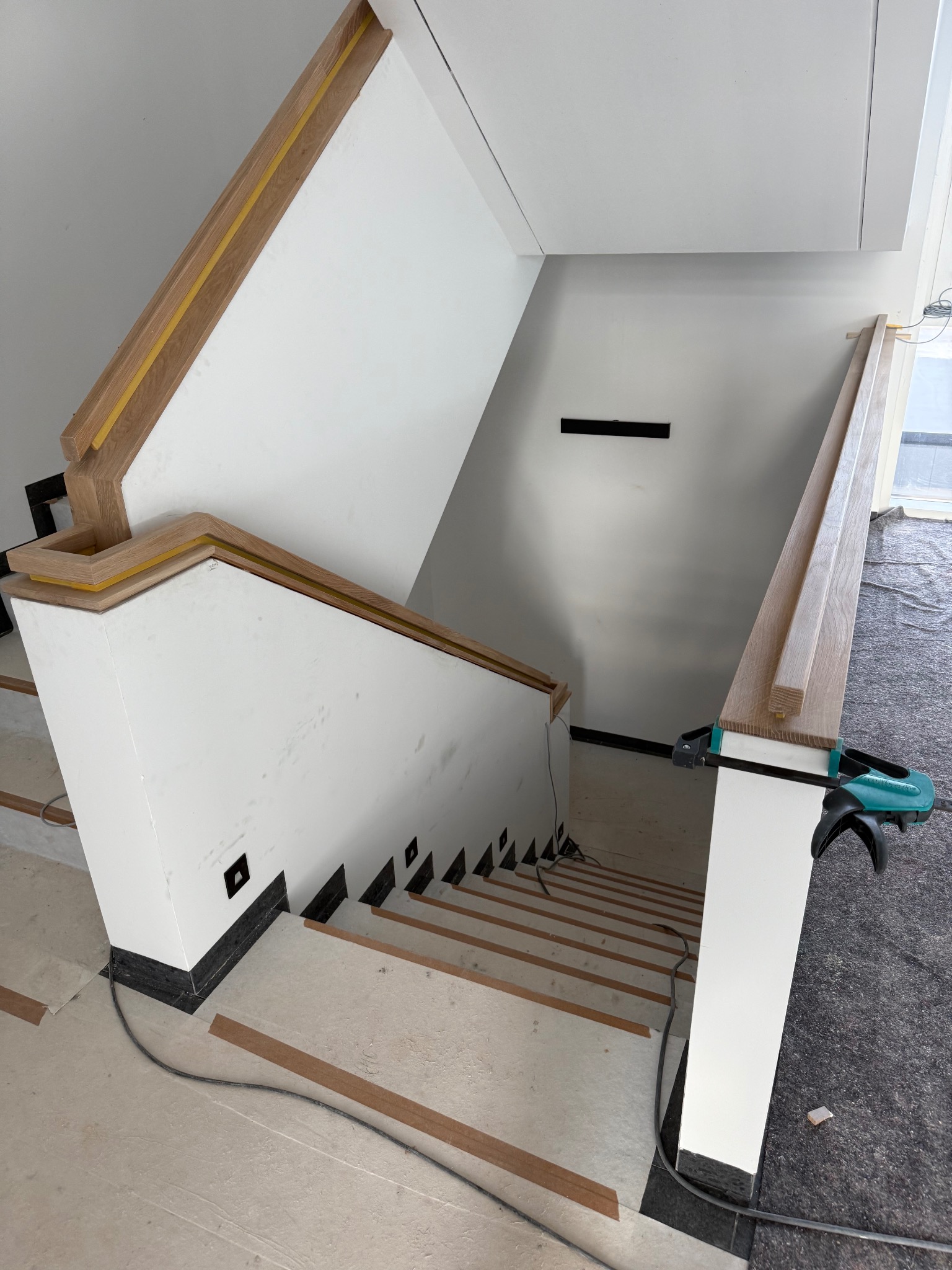 Montaż dębowych schodów z białymi balustradami. Widoczny etap prac z narzędziami. Ujęcie z góry na wnętrze w trakcie wykończenia.