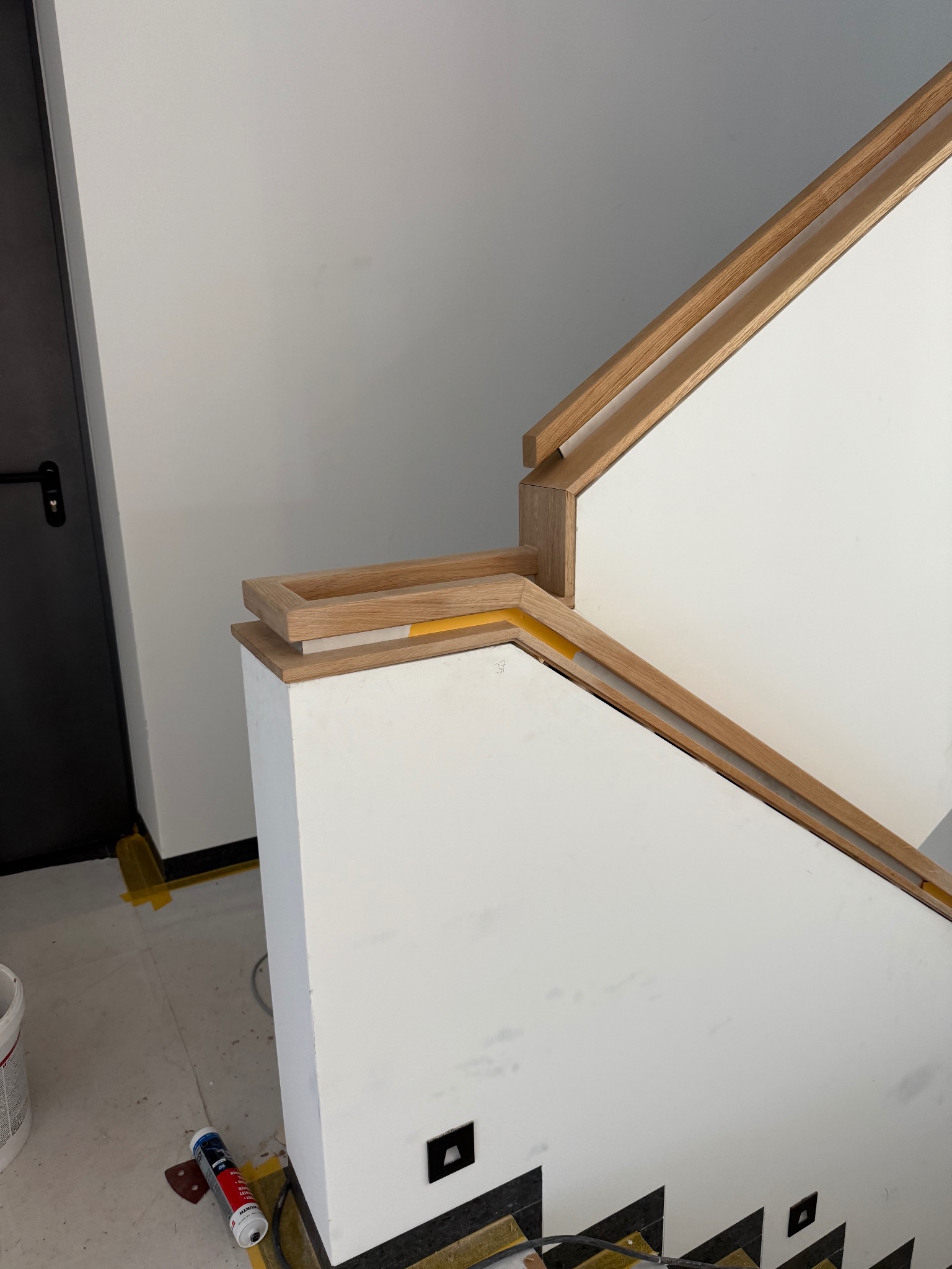 Nowoczesne schody z drewnianą balustradą i białymi panelami. W tle widoczna czarna ściana z drzwiami. Na podłodze narzędzia.