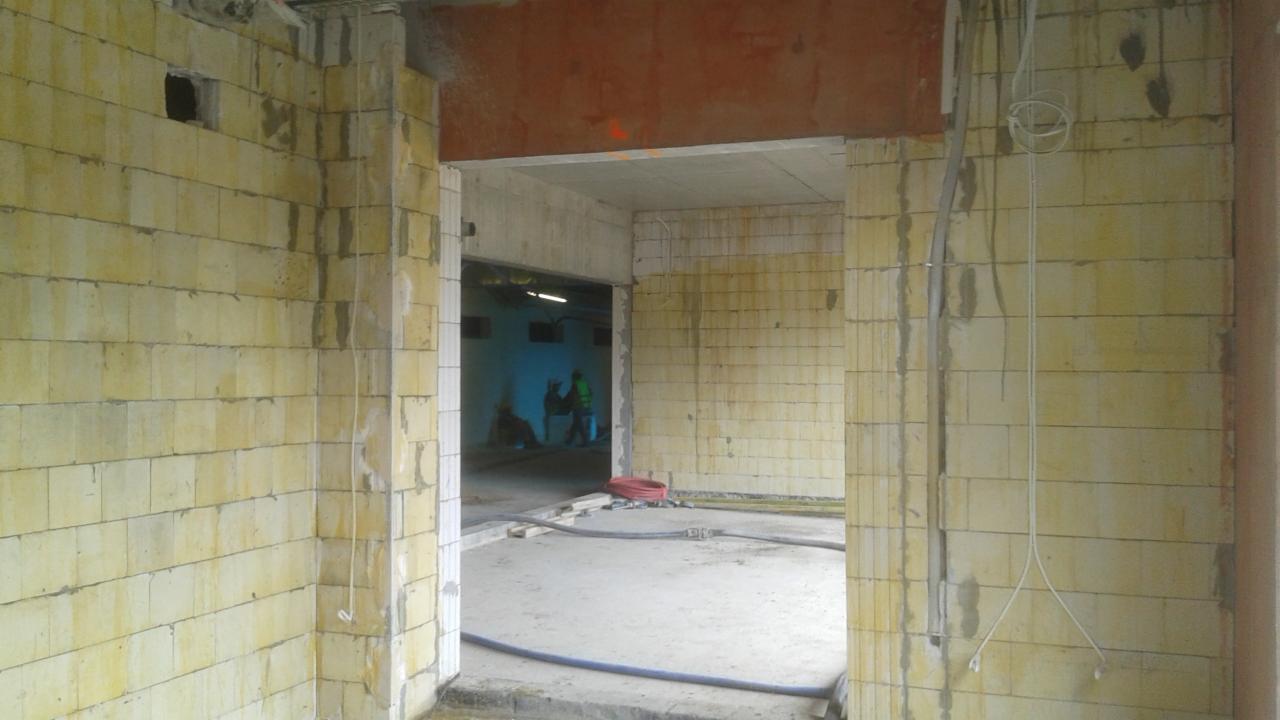 Wnętrze budynku w trakcie budowy, widoczne ściany z bloczków, betonowa podłoga, przewody elektryczne zwisające ze ścian, w tle osoby w odblaskowych kamizelkach.