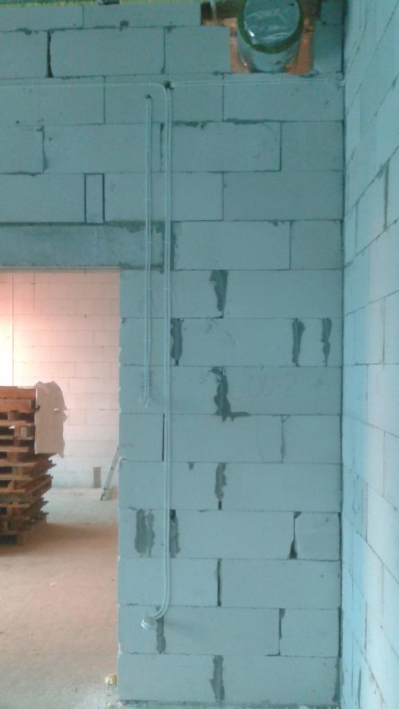 Biała ściana z betonu komórkowego, pionowo zamontowana rurka instalacyjna z białego tworzywa, widoczny fragment puszki instalacyjnej pod sufitem i palety w tle.