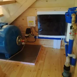 Niebieski zbiornik hydroforowy, filtr wody z niebieskimi elementami i skrzynka elektryczna zamontowane w drewnianej zabudowie pod schodami.