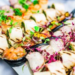 Niku Bowling Monika Gościniak - Elegancko zaaranżowana taca z różnorodnymi przekąskami cateringowymi, w tym roladki z grilla, mini tartaletki i kawałki wędzonego łososia, udekorowane świeżymi ziołami i jadalnymi kwiatami...